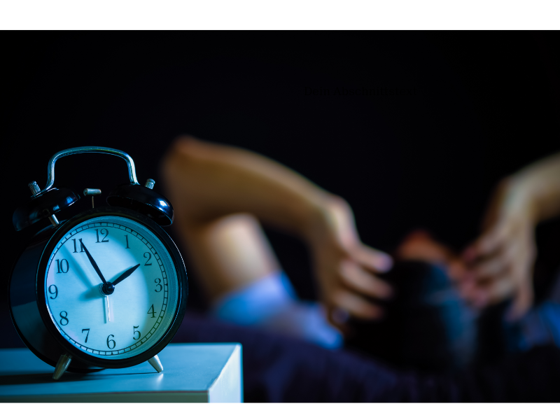 Ein Wecker, dahinter liegt ein Mensch im Bett mit über den Kopf zusammengeschlagenen Händen