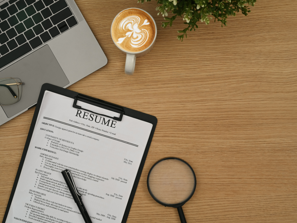 ein Bild von einer Bewerbung vor einem Laptop. Daneben steht ein Kaffee und liegt eine Lupe 