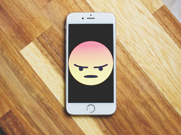 Handy auf einem Holzboden. Auf dem Handy-Bildschirm sieht man ein wütendes Emoji