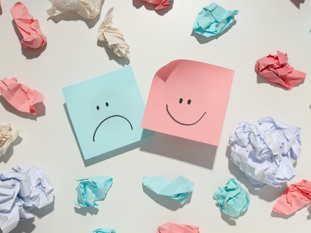 viele kleine zerknüllte Post-Its auf einem weißen Hintergrund. In der Mitte sind zwei Post-Its in blau und rot auf denen jeweils ein trauriger und ein glücklicher Smiley zu sehen ist