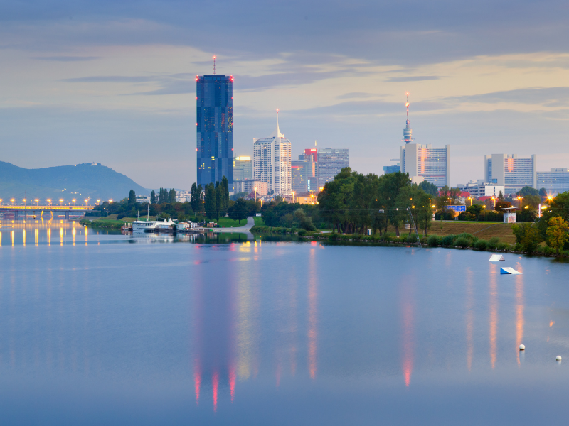 Wiener Skyline an der Donau bei Abenddämmerung mit dem markanten DC Tower 1 und Lichtern, die sich im Fluss spiegeln.