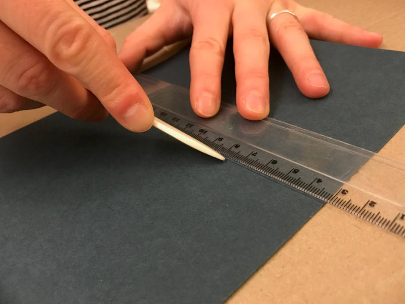 Mit einem Falzbein und einem Lineal wird eine Linie auf einem Papier gezogen.