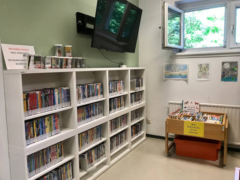 4x4 Regalreihen gefüllt mit DVDs, darüber hängt ein Bildschirm. Daneben steht eine Holzkiste mit der Beschriftung "Bilderbücher für die Kleinsten".