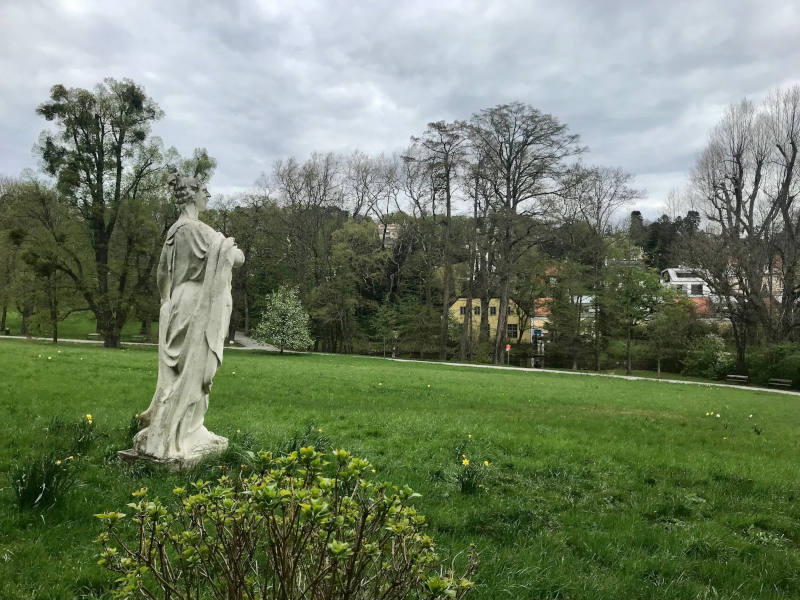 Park, hinter den kahlen Bäumen blitzen Häuser durch. Links sieht man von der Seite eine Attikafigur (Sopran).