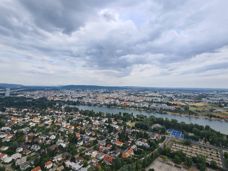Aussicht vom Donauturm: im Vordergrund viele Bäume, vereinzelt Häuser, danach horizontal die Donau, dahinter viele Miniaturhäuser.