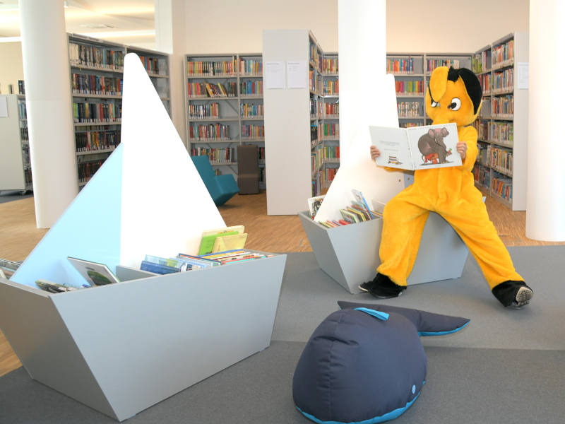 BüchereiAspern Lesefant bei zwei Bücherkisten die wie Schiffe geformt sind und einem Sitzsack-Wal. Im Hintergrund sind Bücherregale.