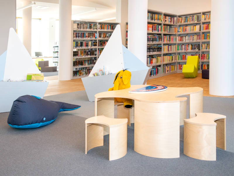 BüchereiAspern Zwei Bücherkisten, die wie Schiffe geformt sind, ein Sitzsack-Wal und Holztisch mit Holzsesseln in Kindergröße mit Lesefant-Plüschi. Im Hintergrund sind Bücherregale.
