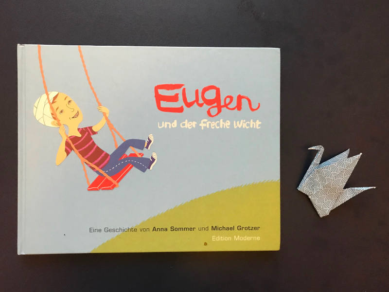 Buch "Eugen und der freche Wicht" von Anna Sommer und Michael Grotzer, daneben ein Papierkranich.