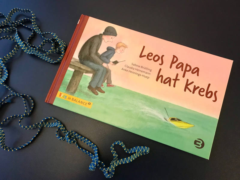 Buch "Leos Papa hat Krebs" von Sabine Brütting und Claudia Heinemann, daneben eine blau-weiße Schnur geschwungen um das halbe Buch.