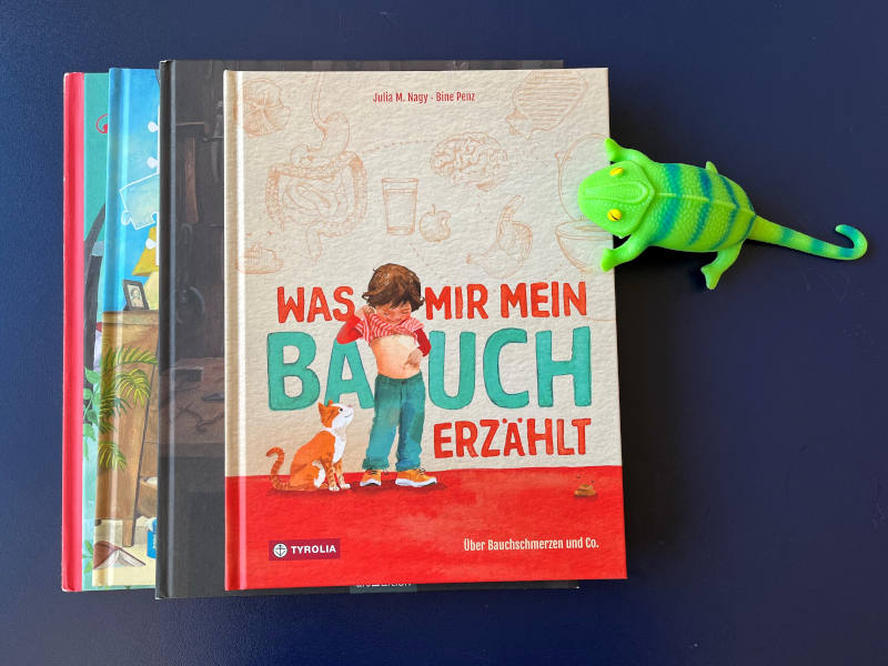 Ein Bücherstapel, ganz oben liegt "Was mir mein Bauch erzählt" von Julia M. Nagy und Bine Penz. Neben dem Bücherstapel ist ein Stoffchamäleon. 