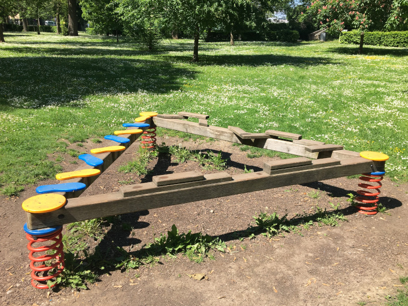 Park in St- Pölten Hammerpark, zu sehen ist ein Dreieck aus Holz mit farbigen, runden Teilen, auf denen Kinder sitzen und balancieren können.