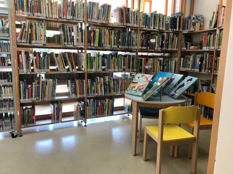 Bücherregale, davor ein Tisch, auf dem 5 Bücher präsentiert werden, mit 3 Holzsesseln.