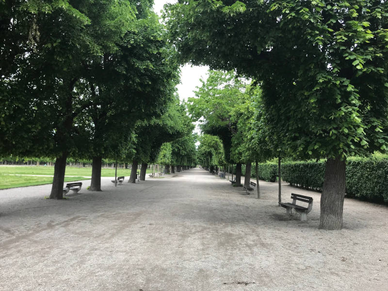 Allee mit Kieselsteinen und Bänken. Rechts davon sind Hecken, links davon Wiese.