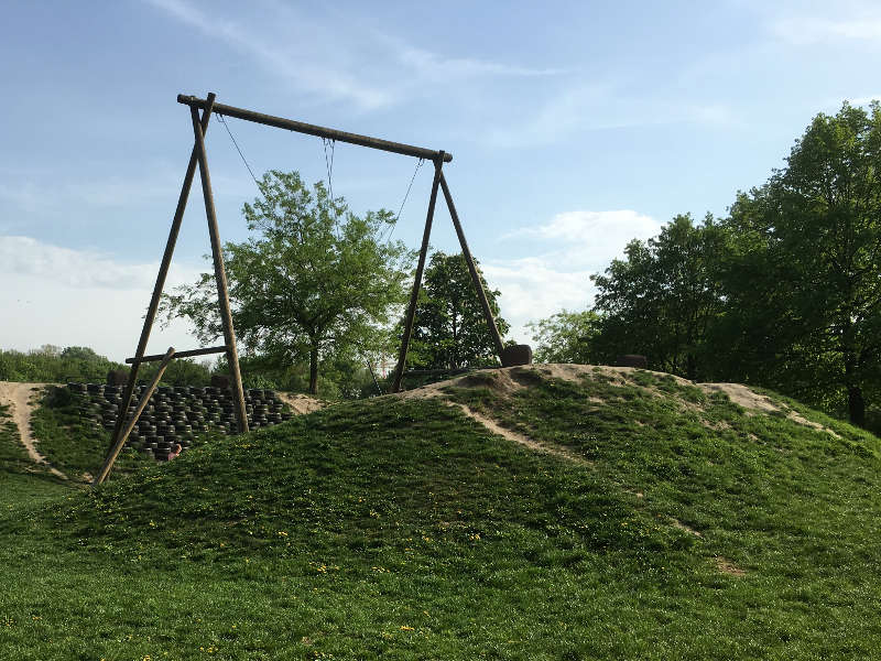 Das Bild zeigt eine große Schaukel auf einem Spielplatz im Freien. Das Kletternetz ist an einem hohen Metallrahmen aufgehängt und spannt sich wie ein Hügel bis zum Boden, der von dichtem grünen Bewuchs umgeben ist. Kinder können über die schrägen Seile nach oben klettern.  Im Hintergrund sind Bäume und offenes Gelände zu sehen, darüber ein heller Himmel. Die Umgebung wirkt weitläufig, ruhig und naturnah – ein Ort, der viel Platz zum Bewegen und Klettern bietet.