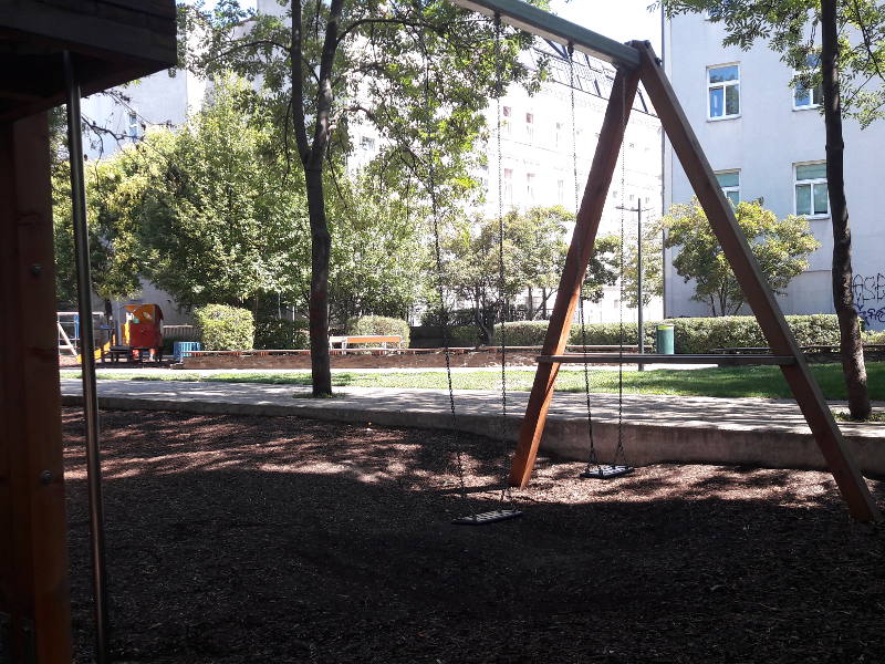 Das Bild zeigt eine einzelne Schaukel auf einem Spielplatz im Freien. Sie hängt an einem schräg stehenden Metallgestell und befindet sich über einem Boden aus Holzschnitzeln, der als Fallschutz dient. Im Hintergrund sind Bäume, parkende Autos und mehrstöckige Wohnhäuser zu sehen, was auf einen Spielplatz in einem städtischen Wohngebiet hinweist. Sonnenlicht fällt durch die Blätter und erzeugt ein warmes, ruhiges Ambiente.