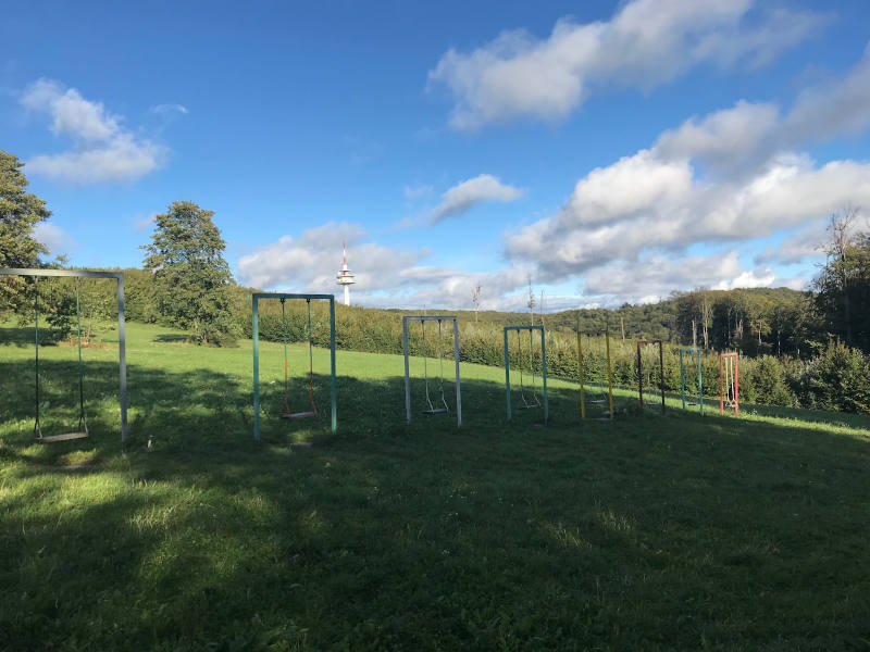 Das Bild zeigt Schaukeln auf einer Wiese im Freien. Die Schaukeln hängen an Metallgestellen und bestehen aus schlichten Sitzbrettern. Sie stehen nebeneinander auf einer grünen Rasenfläche. Im Hintergrund sieht man Bäume, Sträucher und eine leicht hügelige Landschaft unter einem blauen Himmel. Die Umgebung wirkt ruhig, sonnig und naturnah – ein offener Platz, der viel Raum zum Spielen und Schaukeln bietet.