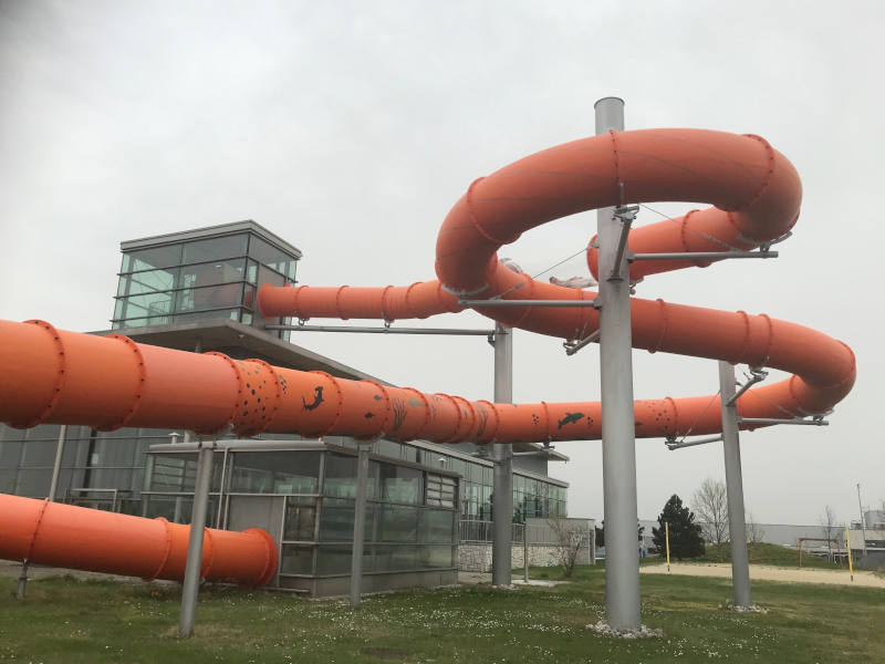 Rote Rutsche von außen, die aus dem ersten Stock von einem Glasgebäude führt, sich hinunter windet und unten wieder ins Gebäude führt. 