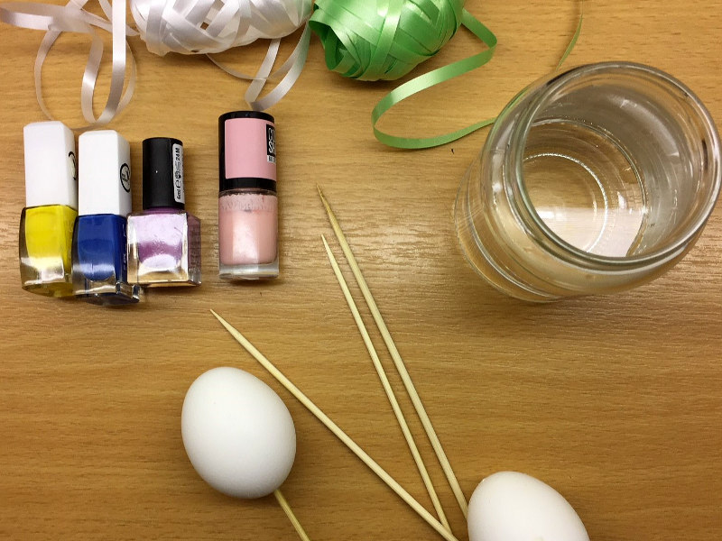 Man sieht einen Tisch auf dem Sachen aufgelegt sind, die man zum Marmorieren“benötigt.  Zu erkennen sind gefärbte Eier, kleine Fläschchen mit Farben, ein Glas mit Flüssigkeit sowie dünne Holzstäbchen. Die Dinge liegen auf einer Holzoberfläche und wirken wie sorgfältig vorbereitetes Zubehör für eine kreative Bastel- oder Färbeaktion.