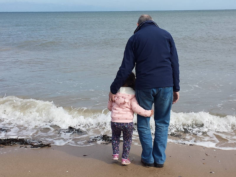 Kind, das sich an einen Erwachsenen schmiegt, vor einem Strand von hinten fotografiert