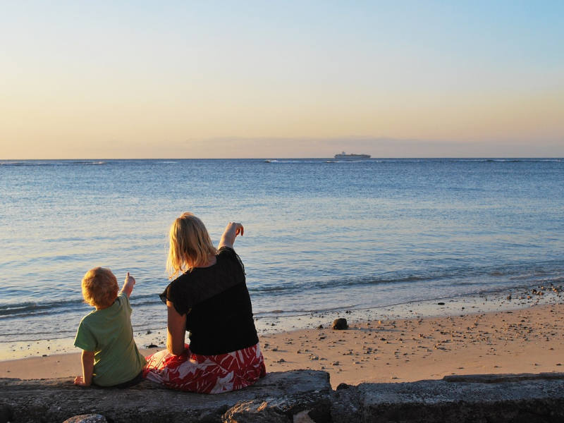 Frau und Kind am Meer sitzend im Sonnenuntergang von hinten