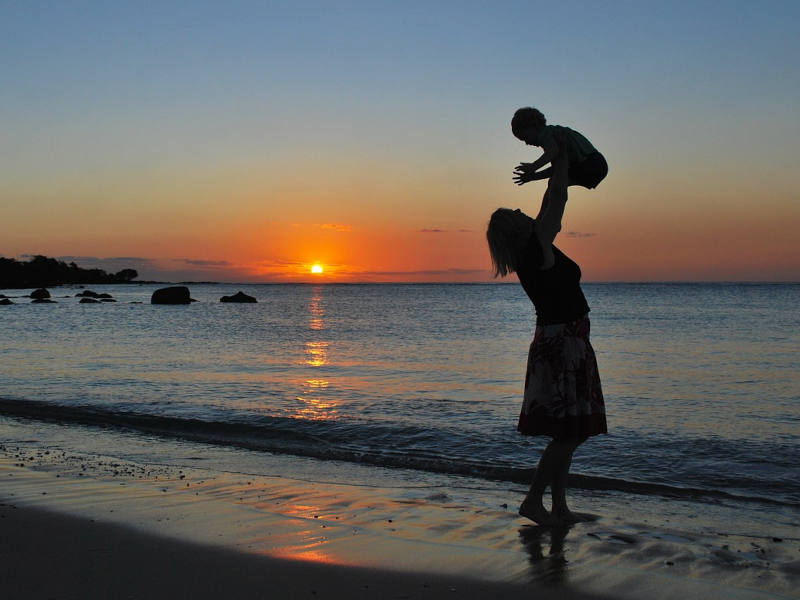 Frau hebt ein Kind in die Höhe vor einem Sonnenuntergang