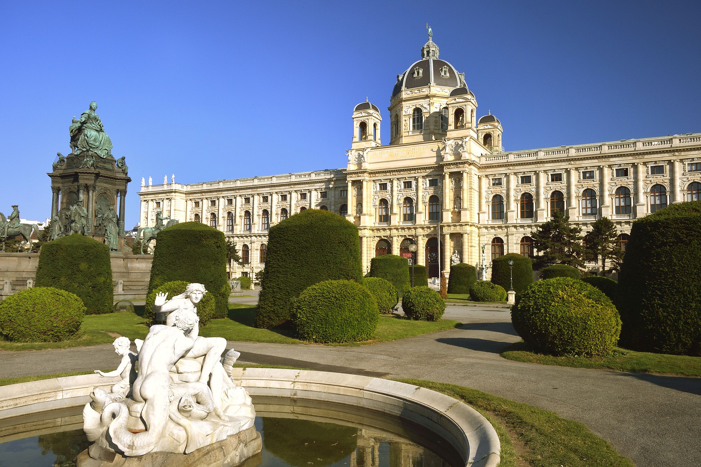 Im Vordergrund ein Brunnen mit Statue, im Hintergrund das Naturhistorische Museum von vorne, dazwischen die Wege und Hecken des Maria-Theresien-Parks. Ganz links die Statue von Maria Theresia.