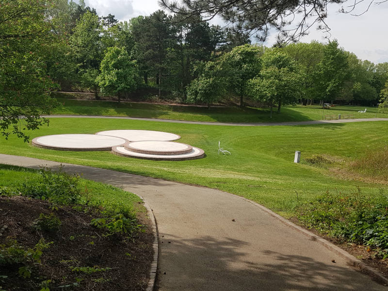 Ein Park mit Bäumen am Außenrand und Wiese in der Mitte. Ein Weg geht gerade und nach links weg. In der Wiese sind vier runde Plattformen.