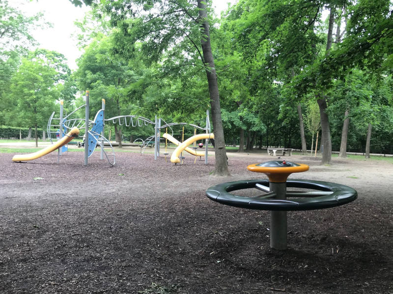 Spielplatz mit einem Metall-Kletterbereich und drei gelbe Rutschen. Im Vordergrund ist ein Karussell für kleinere Kinder zu sehen.