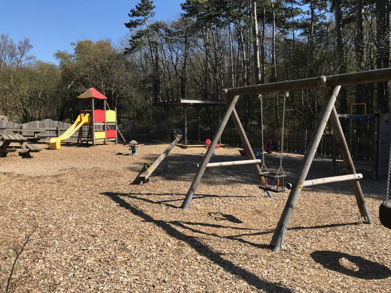 Eine Kinderschaukel im Vordergrund, dahinter zwei Wippen und dahinter ein Turm mit Rutsche, daneben eine Sandkiste. 
