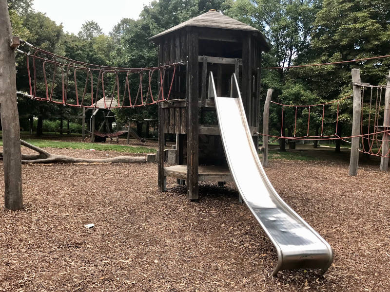 Ein Holzturm mit einer Eisenrutsche. Auf zwei Seiten ist der Holzturm mit einem Seilparkour verbunden.