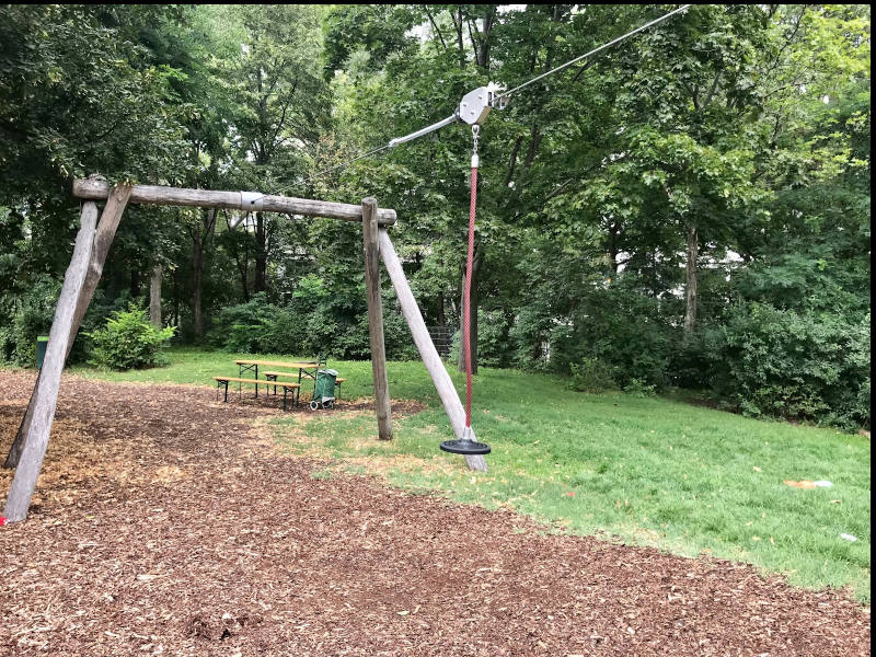 Seilbahn: Zwischen zwei Holzgerüsten mit Höhenunterschied ist ein Seil gespannt. An dem Seil hängt eine beweglicher Sitz, mit dem man das Seil hinab rutschen kann.
