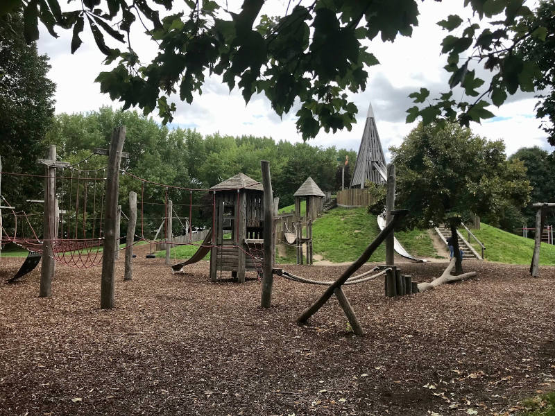 Fläche mit Rindenmulch, im Hintergrund ein Grashügel, mit Rutsche. Davor ein Kletterseilpark aus Holzpfeilern, zwischen denen rote Seile auf verschiedene Arten gespannt sind. 