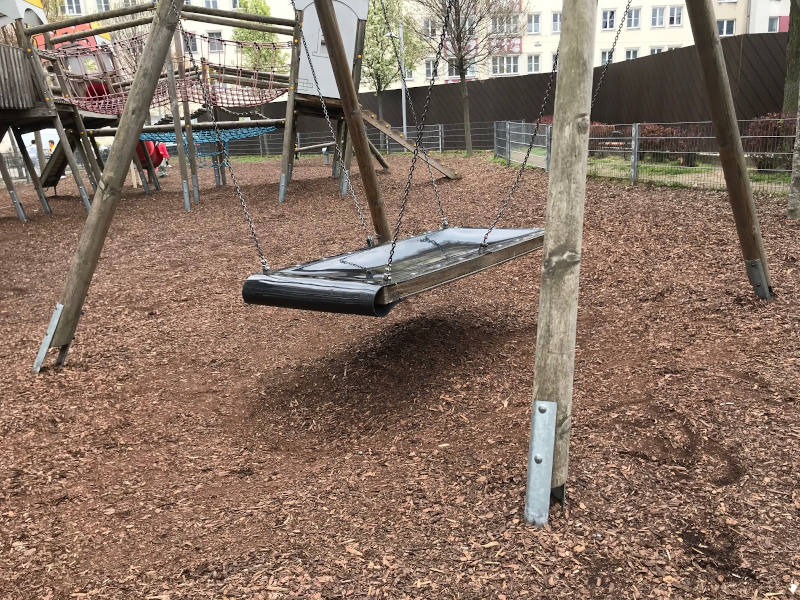 Das Foto zeigt eine Nestschaukel auf einem Spielplatz im Freien. Die runde, schwarze Schaukel hängt an Seilen zwischen zwei Metallpfosten und schwebt über einem Boden aus Holzschnitzeln, die als Fallschutz dienen. Im Hintergrund sind Bäume ohne Blätter, parkende Fahrräder und Gebäude zu sehen, was auf einen städtischen Park oder Innenhof hindeutet. Die Szene wirkt ruhig und herbstlich, und die Schaukel ist im Moment unbenutzt.