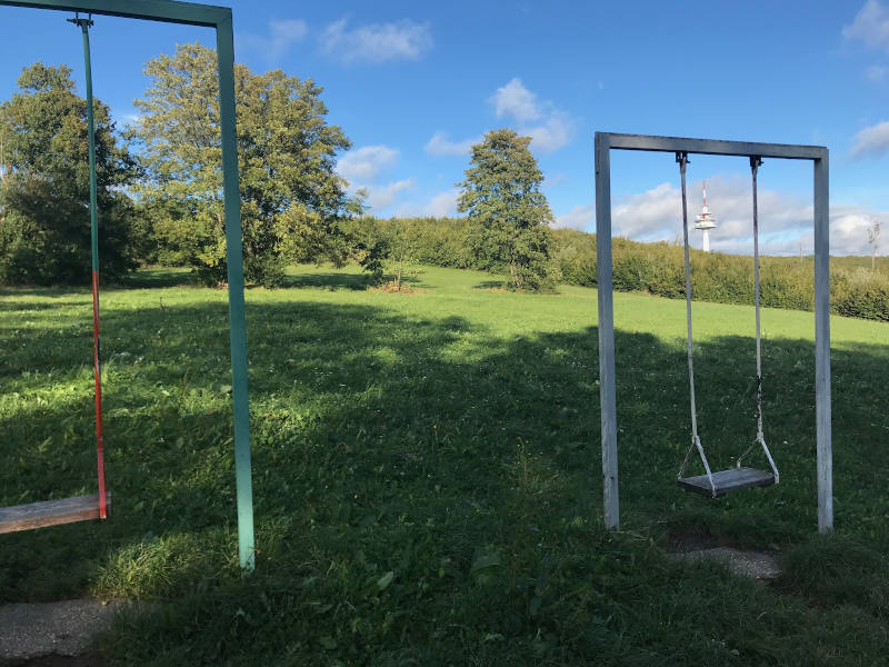 Das Bild zeigt zwei einfache Schaukeln auf einer Wiese im Freien. Die Schaukeln hängen an Metallgestellen und bestehen aus schlichten Sitzbrettern. Sie stehen nebeneinander auf einer grünen Rasenfläche.  Im Hintergrund sieht man Bäume, Sträucher und eine leicht hügelige Landschaft unter einem blauen Himmel. Die Umgebung wirkt ruhig, sonnig und naturnah – ein offener Platz, der viel Raum zum Spielen und Schaukeln bietet.