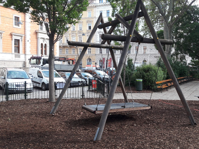 Das Bild zeigt ein großes Klettergerüst aus Holz in Form eines Dreiecks auf einem Spielplatz im Freien. Zwischen den schrägen Holzbalken sind Seile gespannt, an denen Kinder klettern können. Der Boden ist mit Laub und Holzschnitzeln bedeckt. Im Hintergrund sieht man parkende Autos, Fahrräder und mehrstöckige Wohnhäuser, wodurch der Spielplatz in einem städtischen Umfeld liegt. Bäume rahmen die Szene ein und verleihen dem Ort eine herbstliche, ruhige Atmosphäre.