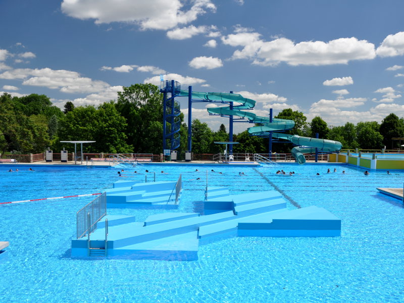 Im Vordergrund ein großes Schwimmbecken mit Podien in der Mitte. Dahinter eine Wendeltreppe, die zu einer blauen Rutsche hoch führt, welche sich dreimal zum Wasser hinab dreht. 