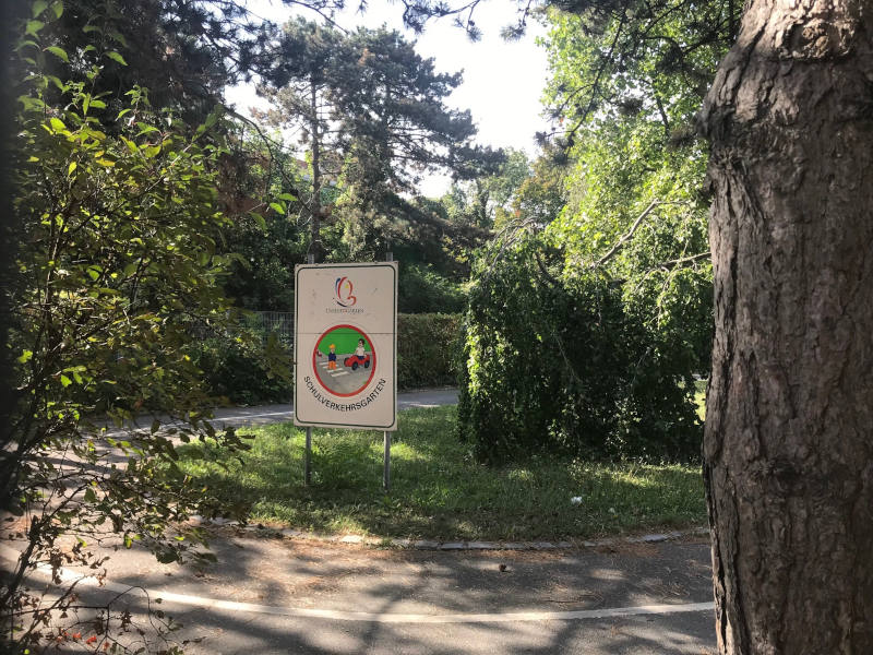 Eine asphaltierte Kurve mitten im Grünen. Auf einer Wiese steht ein Schild mit "Schulverkehrsgarten".