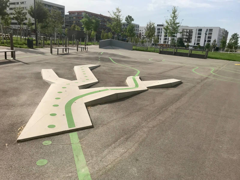 Großer asphaltierter Bereich. Radübungsplatz mit grünen Bodenmarkierungen für verschiedene Trainings. Im Vordergrund eine Erhöhung, die sich in vier Wege abzweigt. Eine gepunktete und eine durchgehende grüne Linie führen über zwei der Abzweigungen.