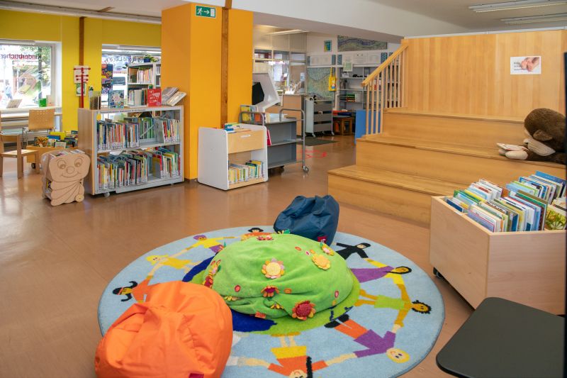 Bunter Teppich mit Sitzsäcken und einer Bücherkiste im Vordergrund, dahinter Holzstiegen und mehrere Bücherregale.