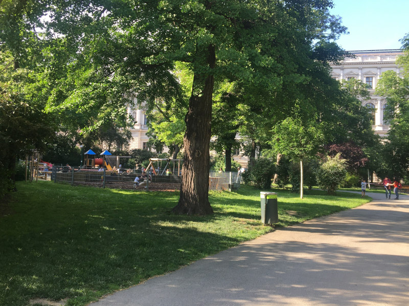 Park mit Weg auf der rechten Seite, Wiese und Baum auf der linken. Im Hintergrund ist ein Spielplatz.