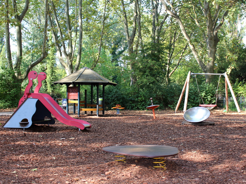 Weiter Blick auf den Spielplatz. Auf der linken Seite ist eine rote Rutsche zu erkennen, im Hintergrund eine Korbschaukel