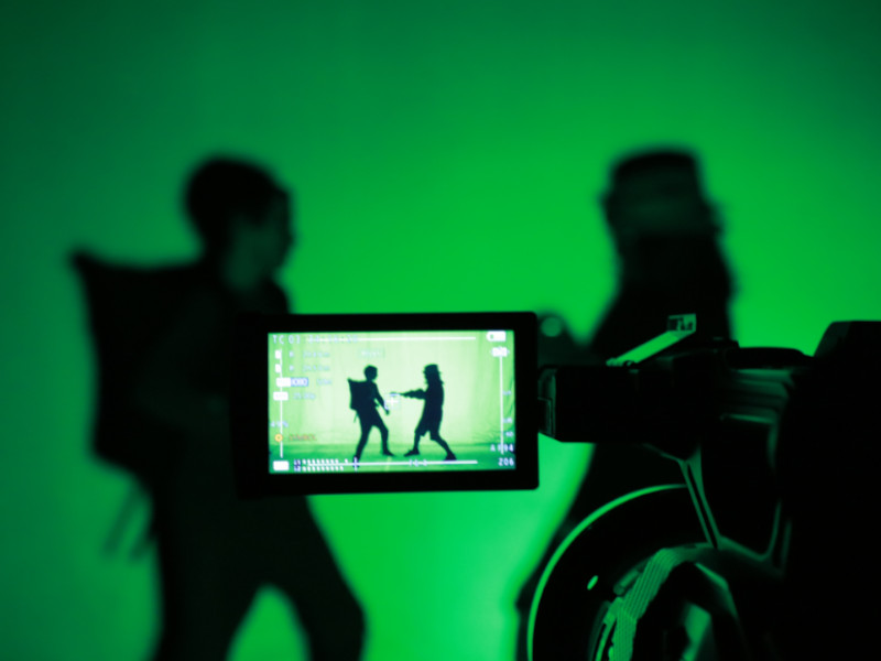 Zwei Personen spielen eine Szene vor einem Greenscreen, während sie von einer Kamera gefilmt werden.