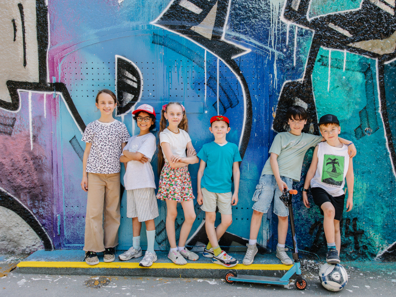 sechs Kinder stehen mit Roller und Fußball vor einer Graffiti-Wand
