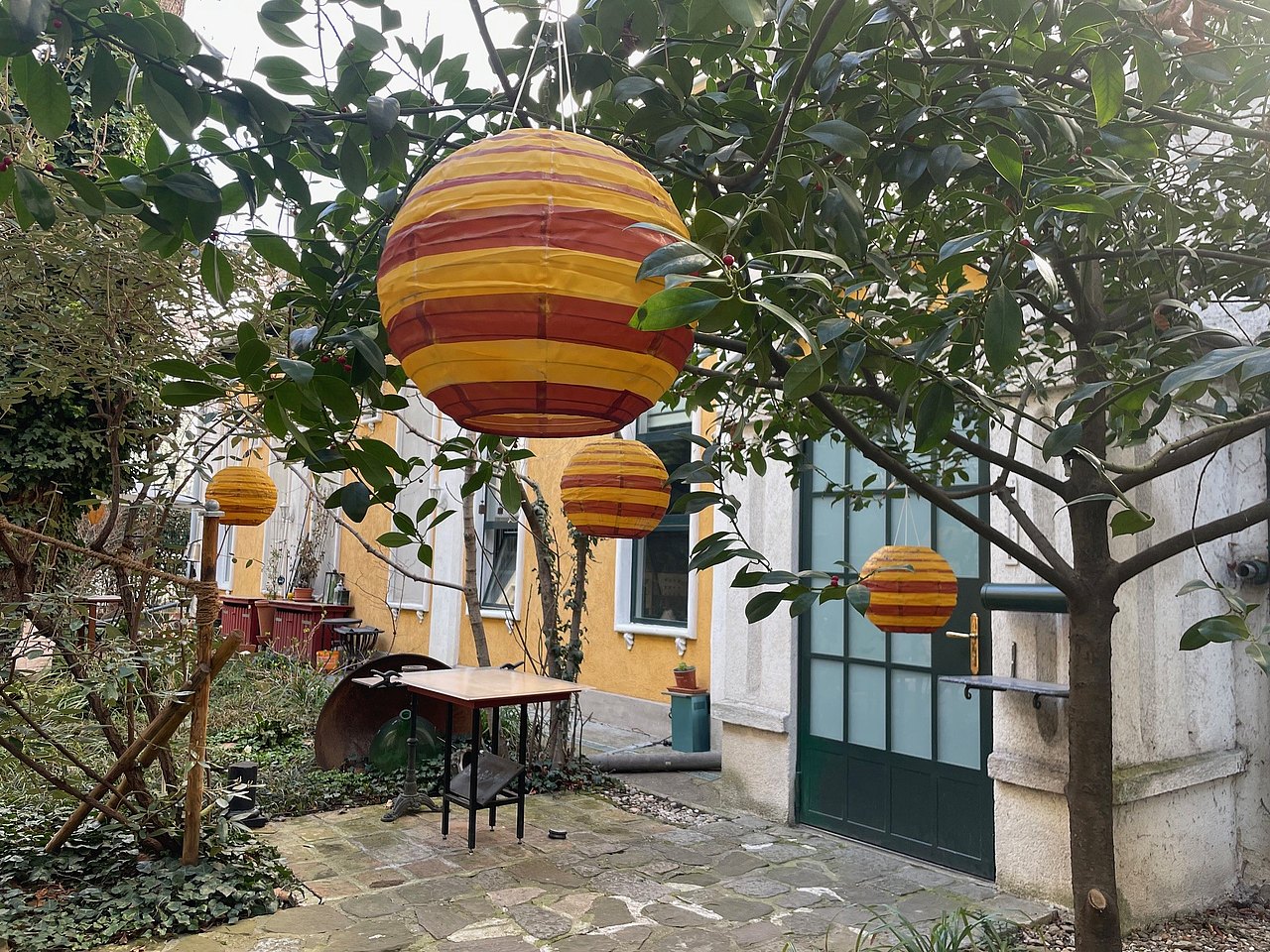 Gelb-rot-gestreifte runde Laternen in einem Innenfof mit kleinem Tisch und Sesseln und Bäumen