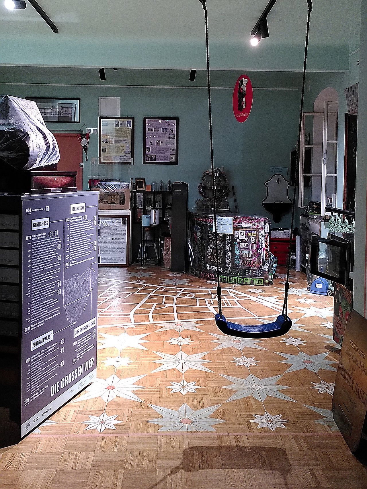 Ausstellungsraum im Bezirksmuseum Neubau mit einer Schaukel im Gang, Infotafeln, einer Theke und Umrisse vom Neubau, umgeben von Sternen am Parkettboden. 