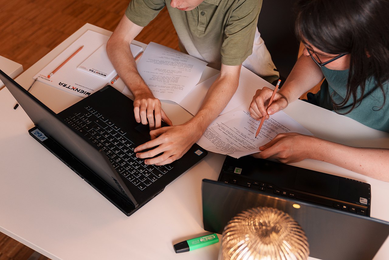 Zwei Personen sitzen an einem Tisch und arbeiten mit Laptops und Zetteln.