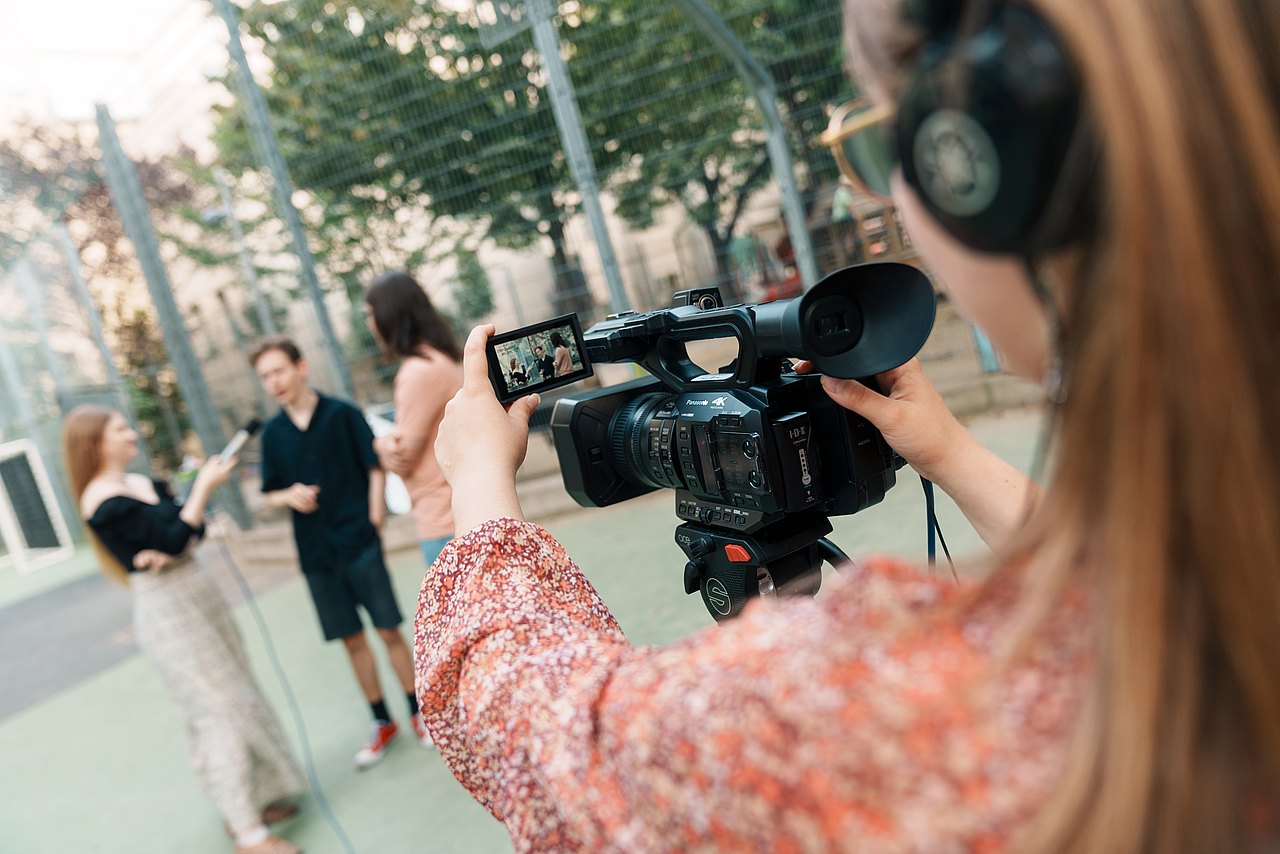 Eine Jugendliche filmt drei andere Jugendliche mit einer Panasonic-Videokamera in einem Park.