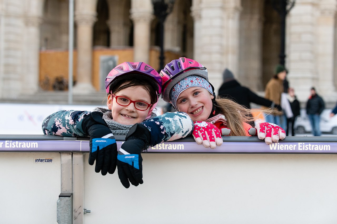 Zwei Mädchen die in Eislaufausrüstung bei der Wand des Eislaufplatzes lehnen