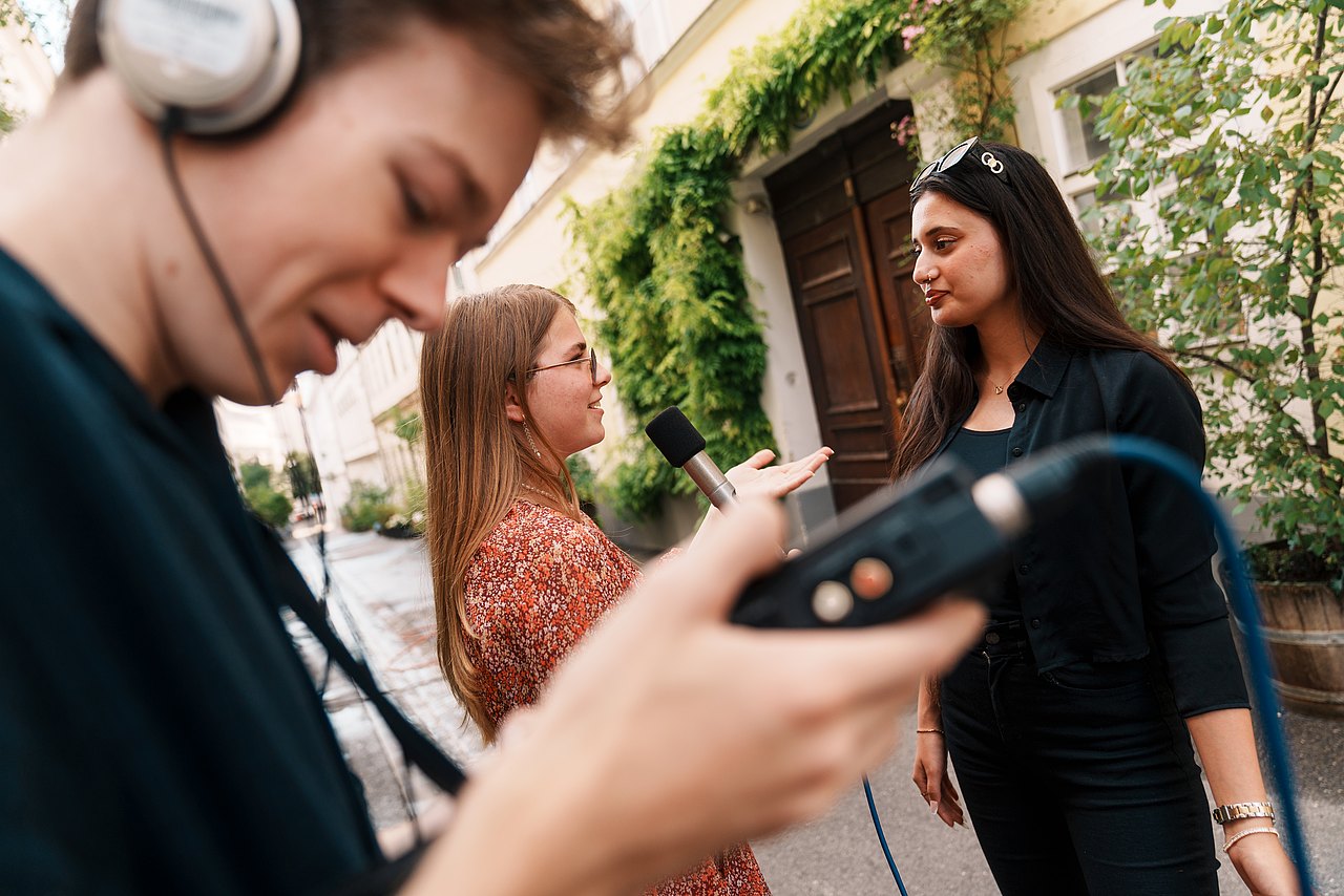 Zwei Jugendliche führen ein Interview mit einem Mikrofon, während ein dritter Jugendlicher Kopfhörer und einen Audio-Recorder trägt 