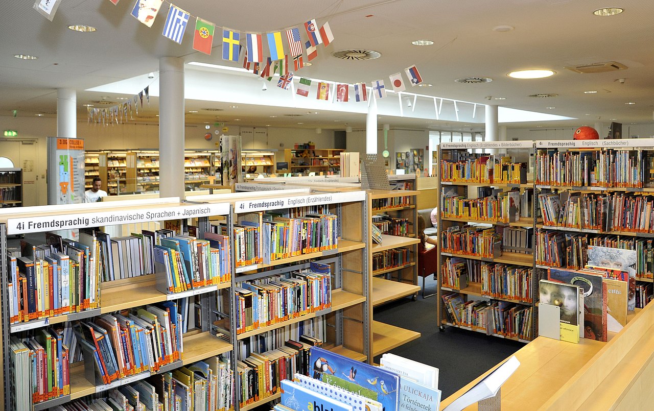 Ein scheinbares Labyrinth aus Bücherkästen. Auf zweien ist "Fremdsprachig skandinavische Sprachen + Japanisch" sowie "Fremdsprachig Englisch (Erzählendes)" zu erkennen. Darüber hängen Girlanden mit Länderflaggen.